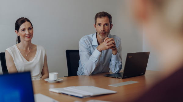 In einer Meetingsituation blicken zwei Personen auf eine dritte im Hintergrund. 